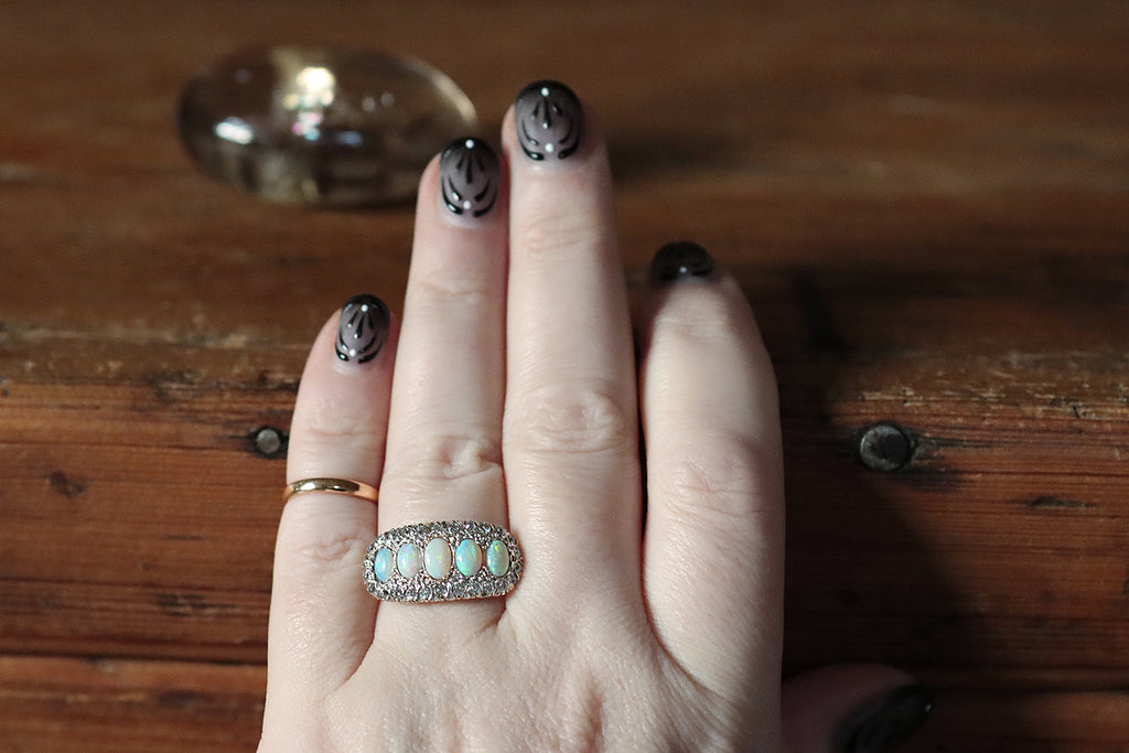 An elegant antique five stone ring, modeled in 14k rose gold, whose primary gems are a series of fiery white opals. These opal cabochons graduate in size across the ring's long, east/west oriented face, which is gently bowed. A border of glittering old cut diamonds, mounted in notched, fishtail settings, surrounds the five opals— the ring's gallery is a decoratively pierced extension of this diamond surround.