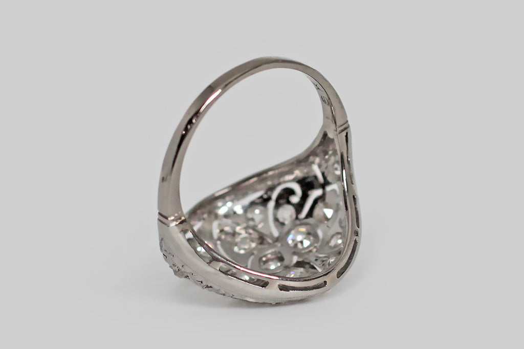 Poor Mouchette | Curated Antique Jewelry, Vintage Jewelry & Engagement Rings | Portland, Oregon | An elegant, early Art Deco diamond cluster ring with Edwardian sensibilities, modeled in 18k white gold with a platinum top. This glamorous, low-profile ring covers the finger like a rounded, tapering shield. It features lovely, hand-pierced decor, that covers the shield-like ring face from edge to edge— the central decor element, here, is a shapely fleur de lis.