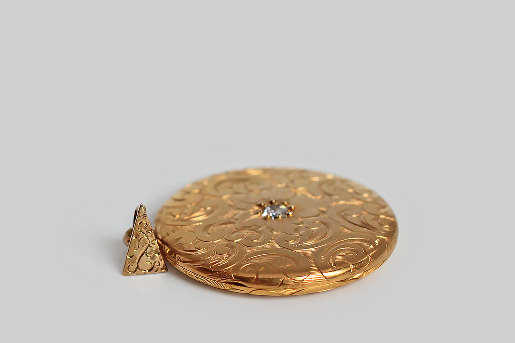 An extra-large, Victorian era locket, modeled in high-color 14k yellow gold, whose face is set with a sparkling 1/3 carat white diamond. The locket is decorated, edge-to-edge, front and back, with ornate, hand-engraved plumes and curls. A plump, languid flower rests amid these swirling forms, at the center of the locket’s face, where the diamond marks its center. This is an especially lovely old locket— the hand engraving is deep, confident, and expressive. 