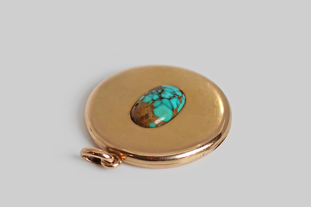 An extra-large, round, Victorian era locket, modeled in 14k rosy yellow gold, whose face is set with a single, large, matrix-webbed turquoise cabochon. This soulful, oval, blue-green gem is highly saturated, and its matrix is reddish-to-golden brown. This locket is clean, minimal, and almost contemporary felling— the earthy turquoise gem seems to float in the simple, gold pool of the locket face. The locket's reverse is hand-engraved with a large monogram, in period script