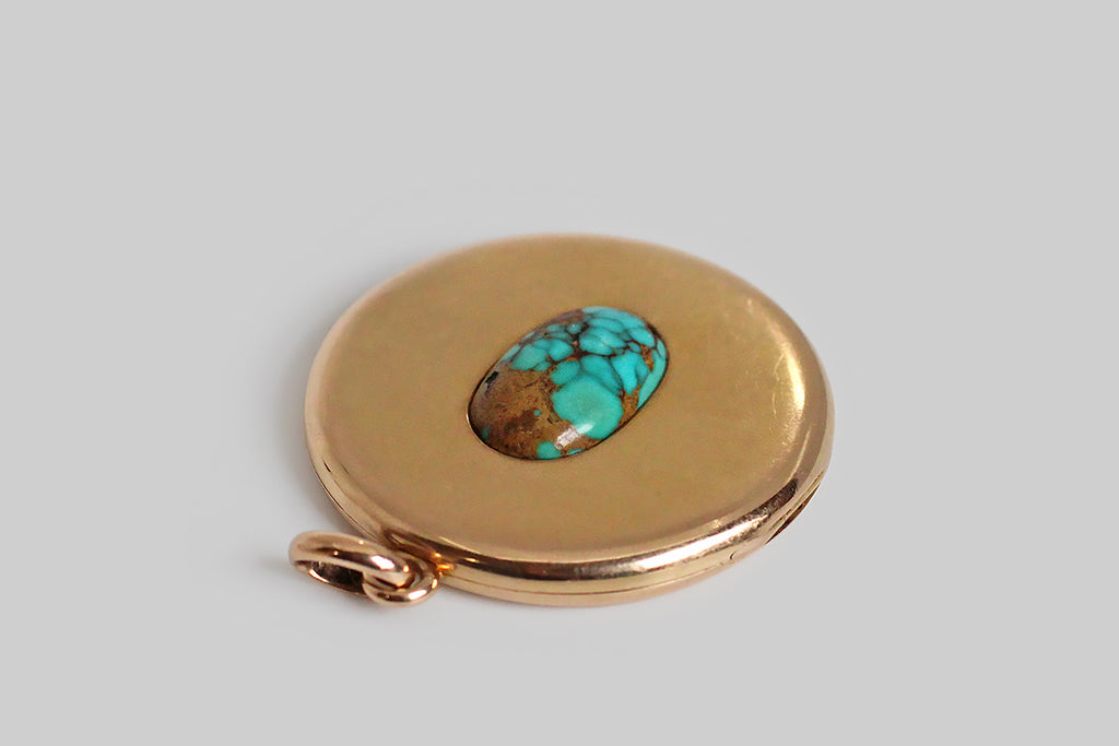 An extra-large, round, Victorian era locket, modeled in 14k rosy yellow gold, whose face is set with a single, large, matrix-webbed turquoise cabochon. This soulful, oval, blue-green gem is highly saturated, and its matrix is reddish-to-golden brown. This locket is clean, minimal, and almost contemporary felling— the earthy turquoise gem seems to float in the simple, gold pool of the locket face. The locket's reverse is hand-engraved with a large monogram, in period script