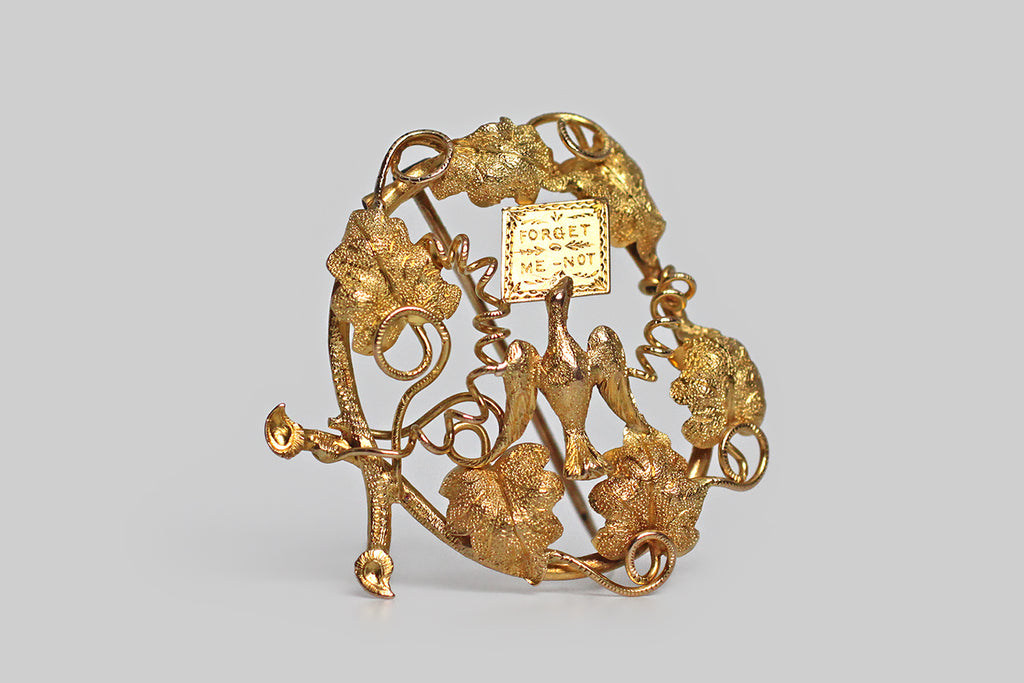 A large, Georgian-era brooch, modeled in 14k yellow gold, whose subject (a dove in flight, bearing a letter) is depicted among grapevines, grape leaves, and tendrils. Our feathered friend's message is a sentimental one— "forget me not." This brooch is primarily repoussé, which means its many, highly-dimensional elements were formed by hammering a gold sheet into relief from the reverse side. 