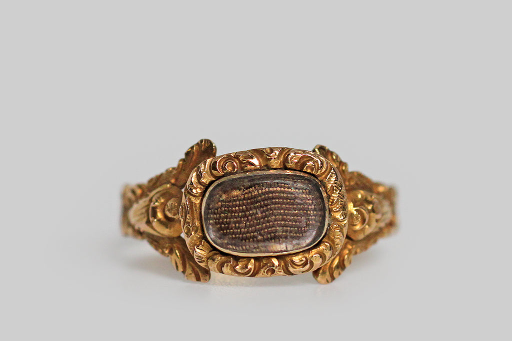A dainty, Georgian-era mourning ring, modeled in high karat gold and decorated with the sumptuous, chased, floral details that are typical of this period. The ring’s highly dimensional shoulders each feature a stylized cabbage rose and a pair of leaves stretching out above it to create the impression of a split shoulder. This shoulder decor is especially voluptuous and creaturely— the ornate bezel that rests betwixt the roses holds tightly plaited locks of human hair, 