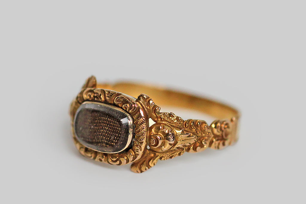 A dainty, Georgian-era mourning ring, modeled in high karat gold and decorated with the sumptuous, chased, floral details that are typical of this period. The ring’s highly dimensional shoulders each feature a stylized cabbage rose and a pair of leaves stretching out above it to create the impression of a split shoulder. This shoulder decor is especially voluptuous and creaturely— the ornate bezel that rests betwixt the roses holds tightly plaited locks of human hair, 