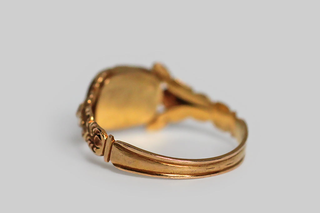 A dainty, Georgian-era mourning ring, modeled in high karat gold and decorated with the sumptuous, chased, floral details that are typical of this period. The ring’s highly dimensional shoulders each feature a stylized cabbage rose and a pair of leaves stretching out above it to create the impression of a split shoulder. This shoulder decor is especially voluptuous and creaturely— the ornate bezel that rests betwixt the roses holds tightly plaited locks of human hair
