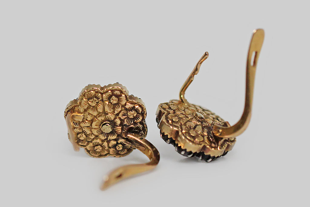 A rare and wonderful pair of dainty, Georgian, button earrings, fabricated in 14k yellow gold and silver, whose rose cut diamonds are arranged to suggest a flower. These glinty old diamonds are mounted in crimped, darkened silver collets, and those collets rest inside gold, flower-shaped, sawtooth-edge bases. The subtly-convex backsides of these earrings are decorated with the most beautiful, deeply-hand-engraved details