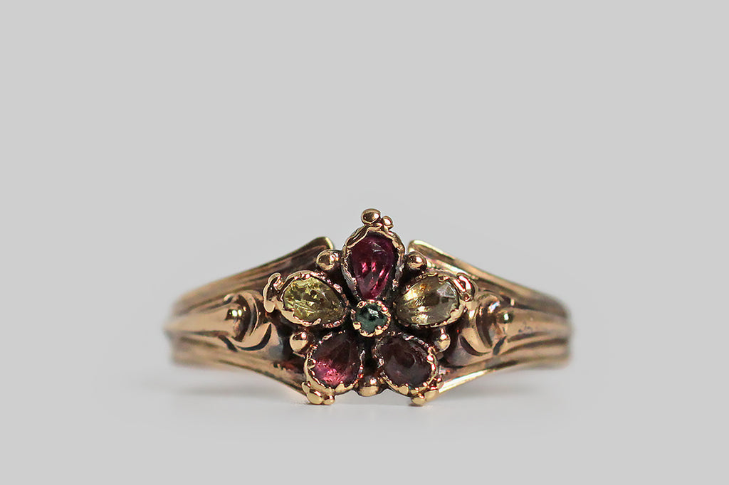 A Georgian-era pansy form ring, modeled in high karat gold, whose petals are set with faceted garnet and chrysoberyl gems. This little flower is highly-dimensional, thanks to the slight pitch of its petals, and the alternating colors of these petals add a loves me, loves me not suggestion to this traditional sweetheart motif. The ring's ornate split shoulders and reeded shank are shapely, unusual, and joined by a delicate shell-like medallion. Pansy is the "think of me" flower, 