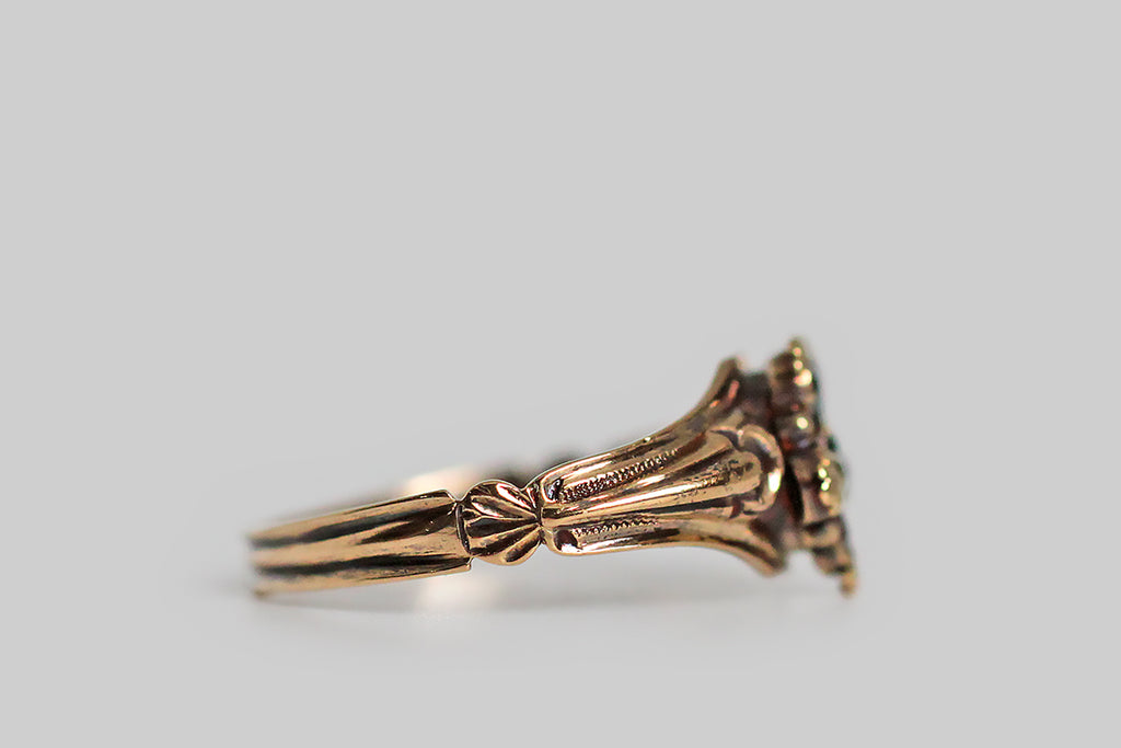 A Georgian-era pansy form ring, modeled in high karat gold, whose petals are set with faceted garnet and chrysoberyl gems. This little flower is highly-dimensional, thanks to the slight pitch of its petals, and the alternating colors of these petals add a loves me, loves me not suggestion to this traditional sweetheart motif. The ring's ornate split shoulders and reeded shank are shapely, unusual, and joined by a delicate shell-like medallion. Pansy is the "think of me" flower, 