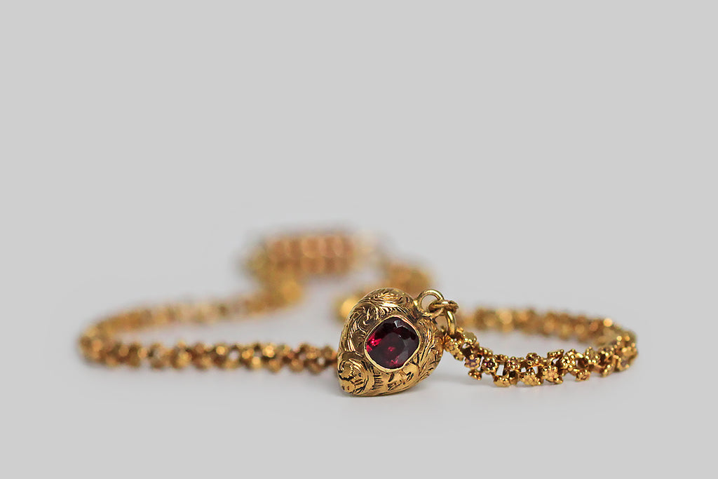 A rare and wonderful Georgian-era bracelet, whose dainty 22k gold links are each decorated with a pair of six-petaled flowers. These high-color, flower-form links encircle the wrist giving the impression of a golden garland. Our garland bracelet also features a miniature heart-shaped locket— the front side of this tiny heart is ornately hand-engraved with baroque plumes, and set with a deep-red garnet.