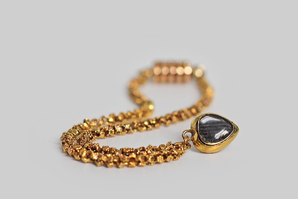 A rare and wonderful Georgian-era bracelet, whose dainty 22k gold links are each decorated with a pair of six-petaled flowers. These high-color, flower-form links encircle the wrist giving the impression of a golden garland. Our garland bracelet also features a miniature heart-shaped locket— the front side of this tiny heart is ornately hand-engraved with baroque plumes, and set with a deep-red garnet.