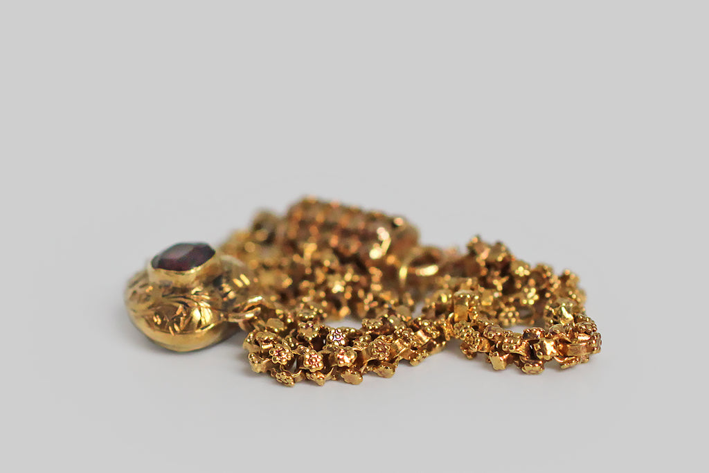 A rare and wonderful Georgian-era bracelet, whose dainty 22k gold links are each decorated with a pair of six-petaled flowers. These high-color, flower-form links encircle the wrist giving the impression of a golden garland. Our garland bracelet also features a miniature heart-shaped locket— the front side of this tiny heart is ornately hand-engraved with baroque plumes, and set with a deep-red garnet.