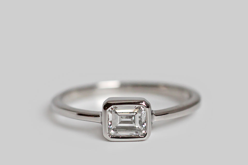 Timeless and elegant, a vintage, half-carat, emerald-cut diamond rests east/west, inside a smooth platinum bezel. This smooth bezel has lovely, crisp lines, and it connects seamlessly to the ring's slender, tapering, square-faced shank. This ring is modern, minimalist, and subtly geometric; its vintage diamond is white (G/H) and very clear (VVS), with an especially large table that creates an icy, hall-of-mirrors effect. This ring is made entirely in platinum