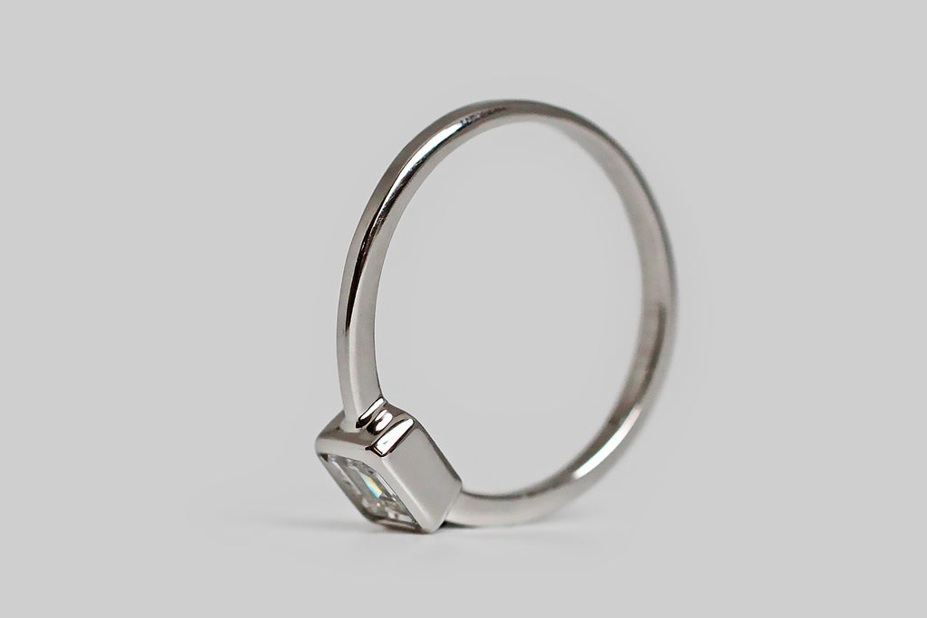 Timeless and elegant, a vintage, half-carat, emerald-cut diamond rests east/west, inside a smooth platinum bezel. This smooth bezel has lovely, crisp lines, and it connects seamlessly to the ring's slender, tapering, square-faced shank. This ring is modern, minimalist, and subtly geometric; its vintage diamond is white (G/H) and very clear (VVS), with an especially large table that creates an icy, hall-of-mirrors effect. This ring is made entirely in platinum