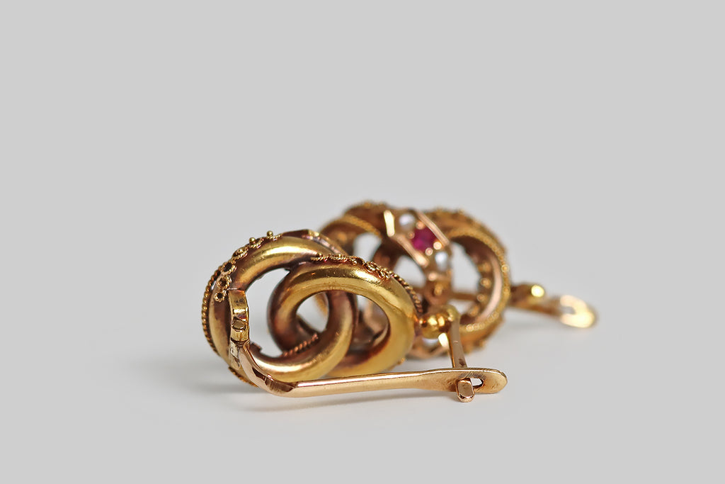 Poor Mouchette | Curated Antique Jewelry, Vintage Jewelry & Engagement Rings | Portland, Oregon | A pair of Victorian-era, Etruscan revival earrings, modeled in 14k rose gold, whose beautiful form resembles a pair of joined rings. These interlocking rings are hollow-form, thick-walled, and fully-dimensional. They are ornately decorated with intricate, twisted wire work, and beads of granulation. Trios of gemstones (ruby and seed pearl) decorate the applied, banner-like seats that rest atop the rings.