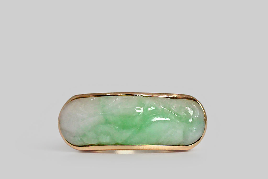 An elegant, large-scale, vintage saddle ring, modeled in 14k yellow gold, featuring a carved, moss in snow, jadeite jade cabochon. This natural, untreated, "A" jade cabochon is held held in a smooth, low-profile bezel— it has been cut on a curve to create the classic saddle shape of the ring, and is set low, so that the cool jade lays against the wearer's skin. This jade cabochon is snow white, with trailing light green figuring. It is carved with soft botanical forms