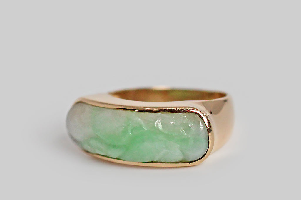 An elegant, large-scale, vintage saddle ring, modeled in 14k yellow gold, featuring a carved, moss in snow, jadeite jade cabochon. This natural, untreated, "A" jade cabochon is held held in a smooth, low-profile bezel— it has been cut on a curve to create the classic saddle shape of the ring, and is set low, so that the cool jade lays against the wearer's skin. This jade cabochon is snow white, with trailing light green figuring. It is carved with soft botanical forms