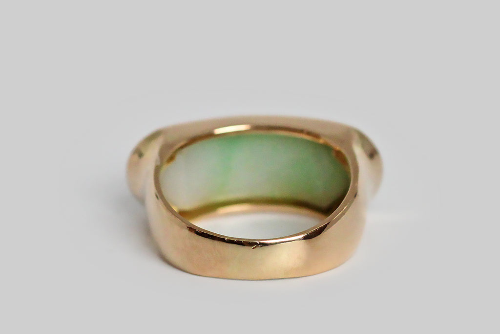An elegant, large-scale, vintage saddle ring, modeled in 14k yellow gold, featuring a carved, moss in snow, jadeite jade cabochon. This natural, untreated, "A" jade cabochon is held held in a smooth, low-profile bezel— it has been cut on a curve to create the classic saddle shape of the ring, and is set low, so that the cool jade lays against the wearer's skin. This jade cabochon is snow white, with trailing light green figuring. It is carved with soft botanical forms