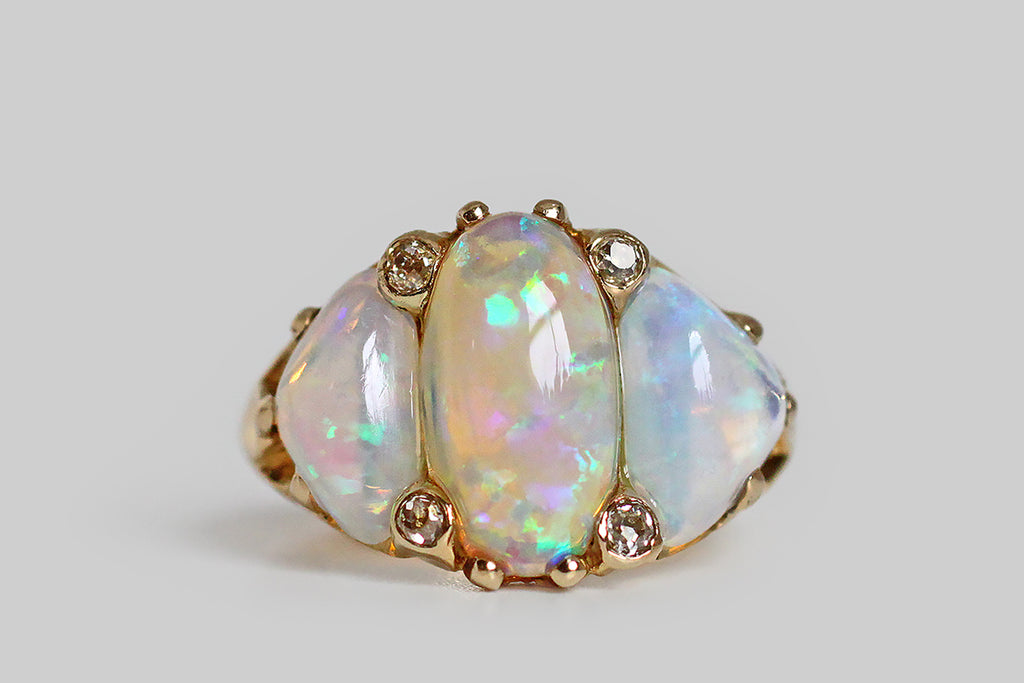 A dazzling, Art Nouveau era ring, modeled in 14k yellow gold and set with a trio of large, dynamic, natural opal cabochons. These crystal opals— a long oval and two bubbly, free-form triangles—  are Australian, and their fiery, rainbow colors (blue, green, pink, purple, and yellow) display in a flag pattern. Between these magical gems, four, small, old mine cut diamonds lay stationed. The organic quality
