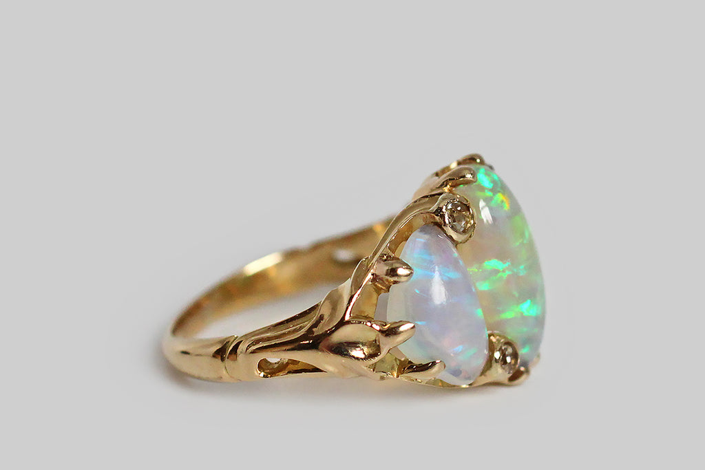 A dazzling, Art Nouveau era ring, modeled in 14k yellow gold and set with a trio of large, dynamic, natural opal cabochons. These crystal opals— a long oval and two bubbly, free-form triangles—  are Australian, and their fiery, rainbow colors (blue, green, pink, purple, and yellow) display in a flag pattern. Between these magical gems, four, small, old mine cut diamonds lay stationed. The organic quality