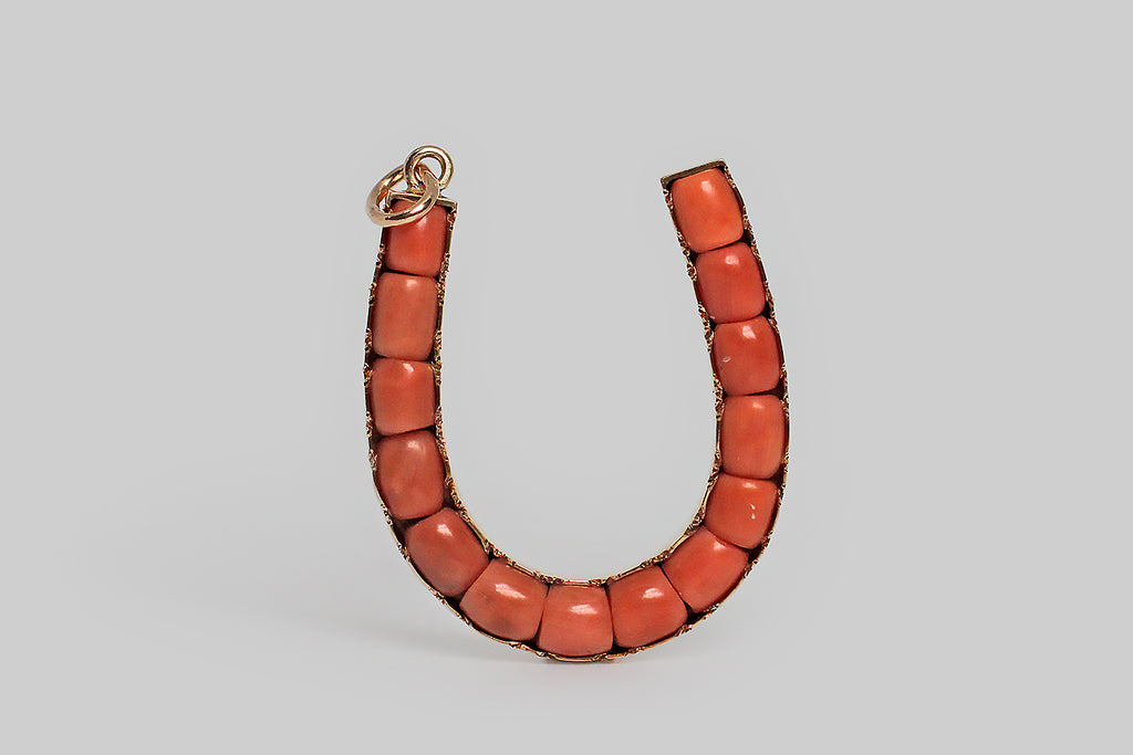 An extra large, Victorian-era, horseshoe pendant, modeled in 14k rose gold, and set with fourteen, natural, untreated coral beads. These Sardinian corals are medium, pink-inflected, orange-red— they are channel-set, end-to-end, into the face of this lucky charm. The horseshoe's frame is hand-fabricated from weighty 14k gold sheet, and its "bezel" is finished with a charming, crimped-and-scalloped, piecrust edge. This is bold and graphic