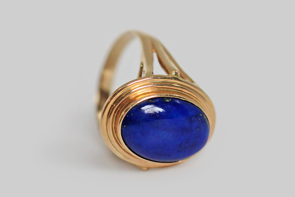 A vibrant, mid-twentieth-century, lapis lazuli ring, modeled in 14k yellow gold, whose large, oval gem is held in a simple, fluted bezel. This ring has split shoulders and an elegant wire work under-gallery that extends the ring's circle-centric vision. The lapis gem is a deeply and evenly saturated blue field, with a few scattered pyrite flecks near the bezel edge— it is every bit as vibrant as it appears, here. 