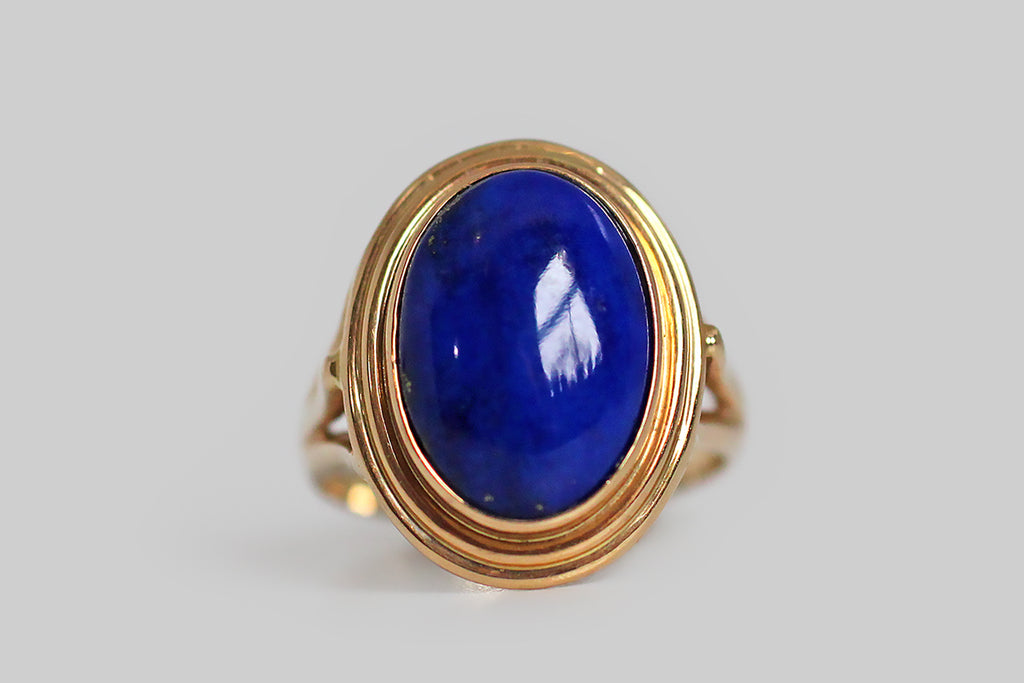 A vibrant, mid-twentieth-century, lapis lazuli ring, modeled in 14k yellow gold, whose large, oval gem is held in a simple, fluted bezel. This ring has split shoulders and an elegant wire work under-gallery that extends the ring's circle-centric vision. The lapis gem is a deeply and evenly saturated blue field, with a few scattered pyrite flecks near the bezel edge— it is every bit as vibrant as it appears, here. 