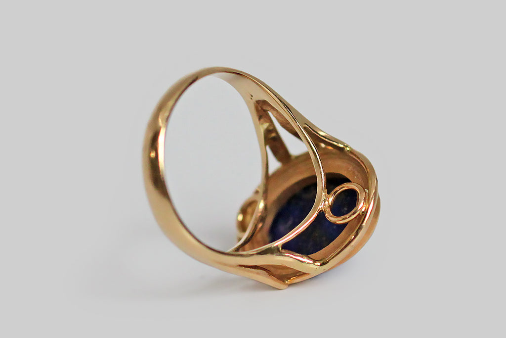 A vibrant, mid-twentieth-century, lapis lazuli ring, modeled in 14k yellow gold, whose large, oval gem is held in a simple, fluted bezel. This ring has split shoulders and an elegant wire work under-gallery that extends the ring's circle-centric vision. The lapis gem is a deeply and evenly saturated blue field, with a few scattered pyrite flecks near the bezel edge— it is every bit as vibrant as it appears, here. 