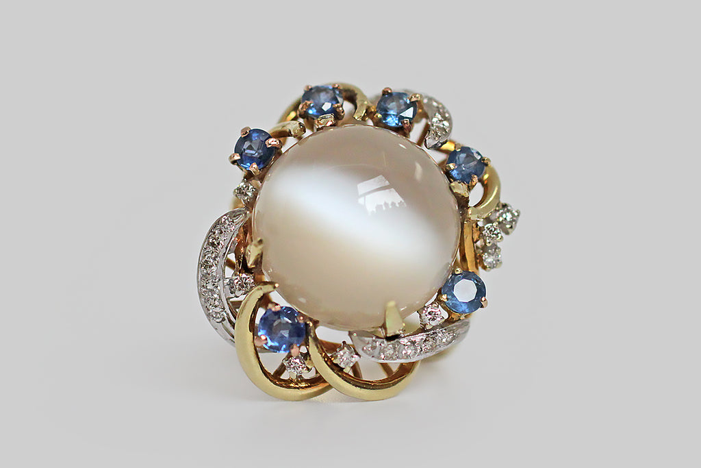 A glorious, mid 20th century, moonstone cocktail ring, made in 14k gold. The ring's big, high-dome, cat’s eye moonstone cabochon displays a strong milk and honey quality, and its adularescence is mesmerizing. This central gem is held in four claw-like prongs; it sits inside an elaborate nest of curling, briar-like, gold forms, some of which are pave-set with small diamonds. 