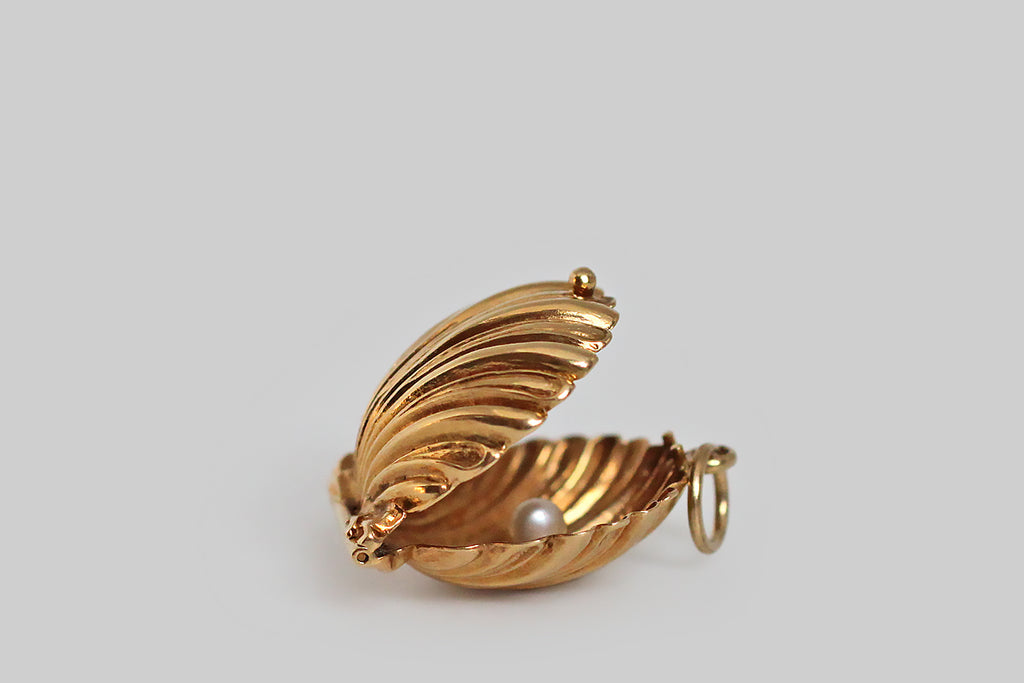 A darling, mid 20th century mechanical charm, modeled in 14k yellow gold, as a seashell that conceals a small white pearl. This little shell has a delicate scalloped form, and the alternating smooth and textured rays of the shell face make it extra pretty. This opens and closes with a latching mechanism— a small bead functions as the push-off. 