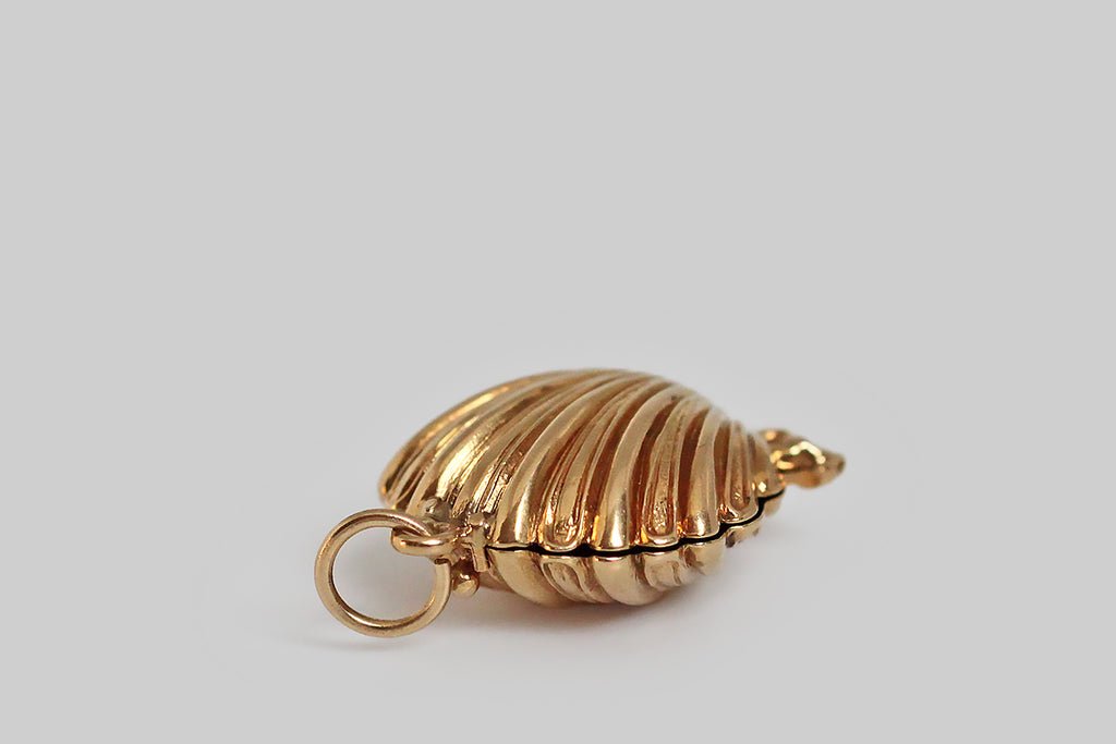 A darling, mid 20th century mechanical charm, modeled in 14k yellow gold, as a seashell that conceals a small white pearl. This little shell has a delicate scalloped form, and the alternating smooth and textured rays of the shell face make it extra pretty. This opens and closes with a latching mechanism— a small bead functions as the push-off. 