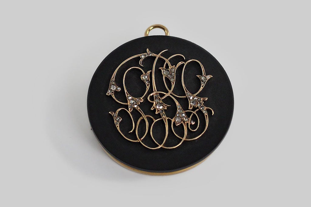 A large, custom made, Georgian era mourning locket with a face of Wedgwood black basalt. This material is very black, truly matte, and subtly porus, like unglazed ceramics. The face is decorated with an elaborate 14k flat-wire monogram, that is embellished with clusters of glinty, rose-cut diamonds. The locket's reverse is a heavy circular mount of 14k gold, encircling a ring of black enamel, encircling a notched ring of gold.