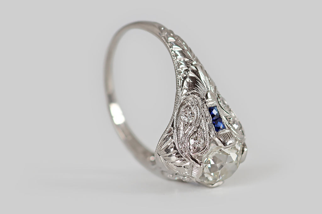 Poor Mouchette | Curated Antique Jewelry, Vintage Jewelry & Engagement Rings | Portland, Oregon | An exceptional Art Deco era engagement ring, modeled in platinum, whose primary gem is a 1.21 carat old mine cushion cut diamond (I/J, SI1). This lively, hand-cut diamond is held in four, tab-style prongs. It is flanked by two pairs of french cut, blue sapphires that grace the ring's elaborate shoulders, reading like dashes astride its large, central gem.