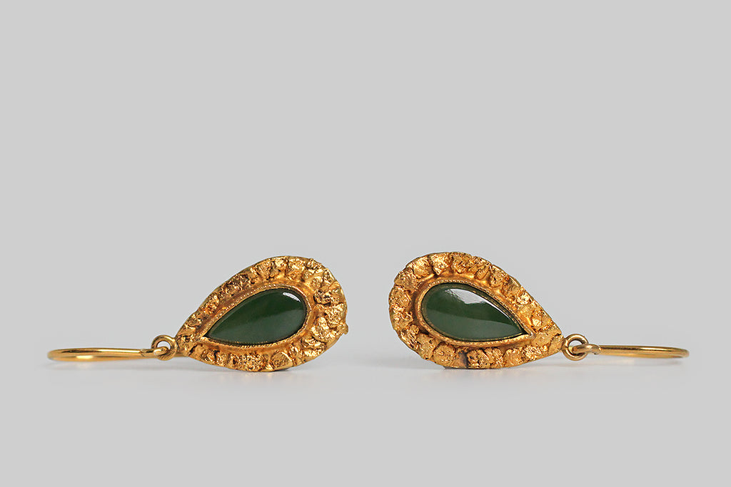 A dainty pair of teardrop-shaped, mid 20th century earrings, whose flat-top, nephrite jade cabochons are surrounded by a deep border of natural gold nuggets. These small, richly-textured placer nuggets are carefully arranged on the earrings' 10k gold base. The teardrops swing below a simple pair of shepherd's hook earwires