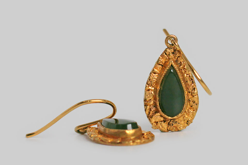 A dainty pair of teardrop-shaped, mid 20th century earrings, whose flat-top, nephrite jade cabochons are surrounded by a deep border of natural gold nuggets. These small, richly-textured placer nuggets are carefully arranged on the earrings' 10k gold base. The teardrops swing below a simple pair of shepherd's hook earwires