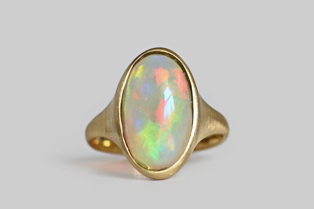 Poor Mouchette | Curated Antique Jewelry, Vintage Jewelry & Engagement Rings | Portland, Oregon | Fiery and changeable, a colorful crystal opal sits, nested, inside a thick-walled, organic bezel, which tapers seamlessly into our ring's integral shank. This elongated oval cabochon is rich with flashes of green, red, yellow, and violet, that present in a brush-stroke pattern, emerging from its translucent, blue-white base and changing as the gem shifts in light. This low-profile