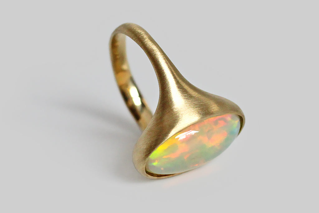 Poor Mouchette | Curated Antique Jewelry, Vintage Jewelry & Engagement Rings | Portland, Oregon | Fiery and changeable, a colorful crystal opal sits, nested, inside a thick-walled, organic bezel, which tapers seamlessly into our ring's integral shank. This elongated oval cabochon is rich with flashes of green, red, yellow, and violet, that present in a brush-stroke pattern, emerging from its translucent, blue-white base and changing as the gem shifts in light. This low-profile