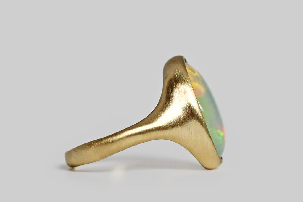 Poor Mouchette | Curated Antique Jewelry, Vintage Jewelry & Engagement Rings | Portland, Oregon | Fiery and changeable, a colorful crystal opal sits, nested, inside a thick-walled, organic bezel, which tapers seamlessly into our ring's integral shank. This elongated oval cabochon is rich with flashes of green, red, yellow, and violet, that present in a brush-stroke pattern, emerging from its translucent, blue-white base and changing as the gem shifts in light. This low-profile
