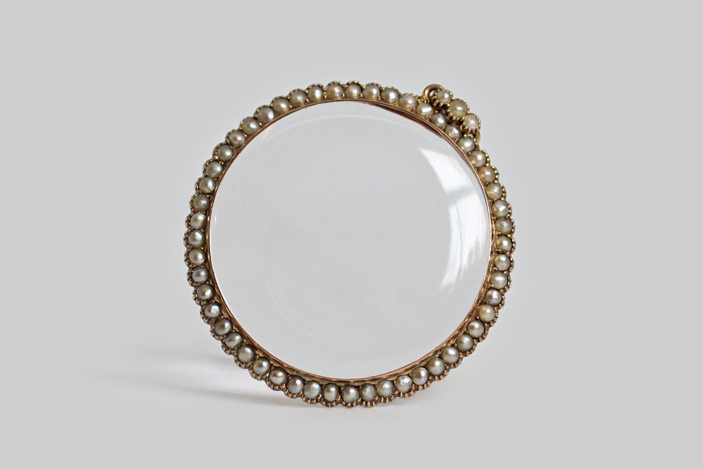 An antique gold locket, made on a grand scale, with a magnifying lens, and a delicate halo of fifty-two, collet-set seed pearls. This airy darling opens easily at the base of the backside, which nests snugly inside the bezel of the front-piece. The locket's large bail is set with three further pearls. This locket is all original. The design is slim and feminine, a perfect frame for whatever treasure is placed inside— a lock of hair, a photograph, a scrap of fabric, a handful of diamonds or colored gems. 