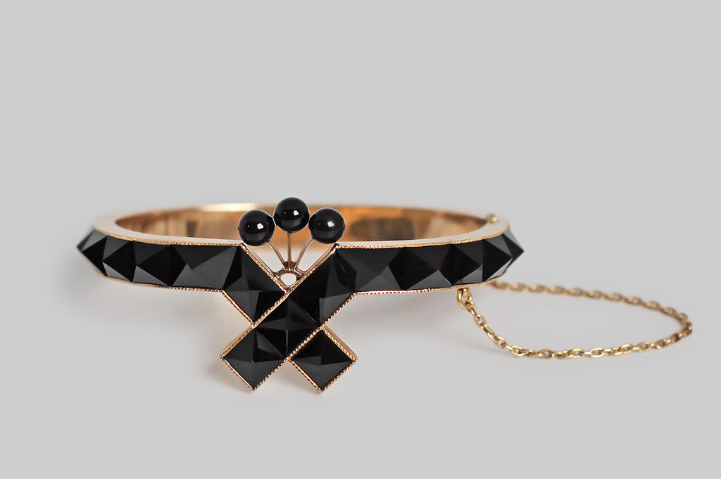 A striking Victorian-era hinged bangle, modeled in 14k rose gold to resemble a mourning ribbon, whose face is set with a series of crisp jet (RI confirmed) pyramids. This crossed ribbon is crowned with a plume-like group of three jet spheres; the backside of the bangle is inlaid with black enamel. 