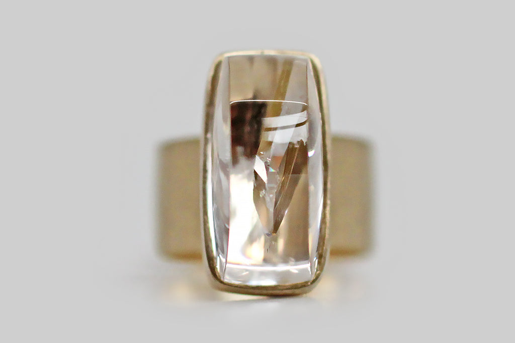 A big, mystical, modernist ring, modeled in 14k yellow gold, that features a rare, axe-head-shaped quartz-in-quartz cabochon. The "mother" quartz, here, is especially fine and optically clear— it holds within it a beautifully formed, iridescent, quartz crystal point that grew into the clear host. This inspiring specimen is mounted north to south, and is seated in a full bezel— it perches atop a shapely, flat pier that connects the ring's head to its wide, flat band. 