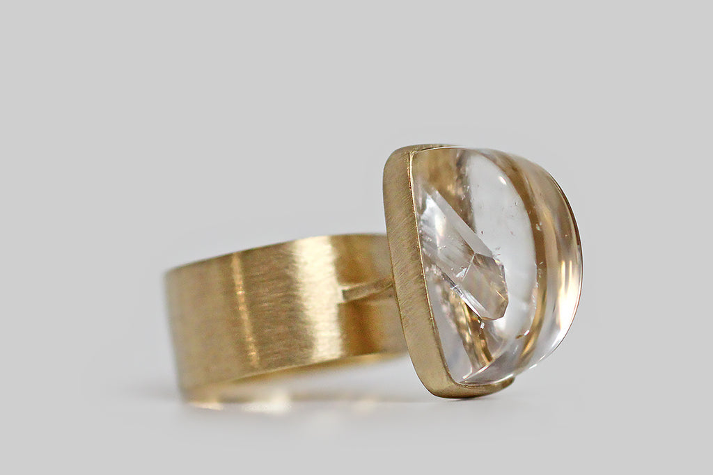 A big, mystical, modernist ring, modeled in 14k yellow gold, that features a rare, axe-head-shaped quartz-in-quartz cabochon. The "mother" quartz, here, is especially fine and optically clear— it holds within it a beautifully formed, iridescent, quartz crystal point that grew into the clear host. This inspiring specimen is mounted north to south, and is seated in a full bezel— it perches atop a shapely, flat pier that connects the ring's head to its wide, flat band. 