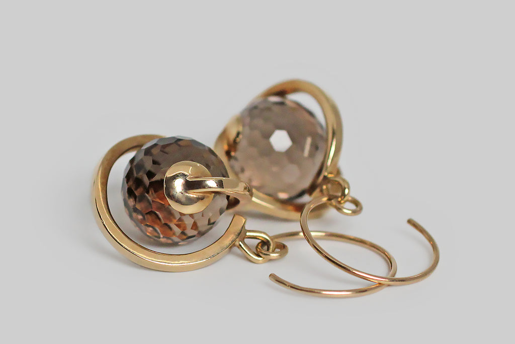 An unusual pair of kinetic earrings, modeled in 14k yellow gold, whose smoky quartz spheres are covered with many, small, hexagon-shaped facets, for extreme light play. These sparkling quartz gems turn freely inside the hand-fabricated, orrery-like mounts, which swing below their earwires. These earrings read like sparkling, ringed planets. The stylized, round, French hooks reflect that spinning aesthetic. These are fun, and eye-catching, with a great warm-neutral color palate.