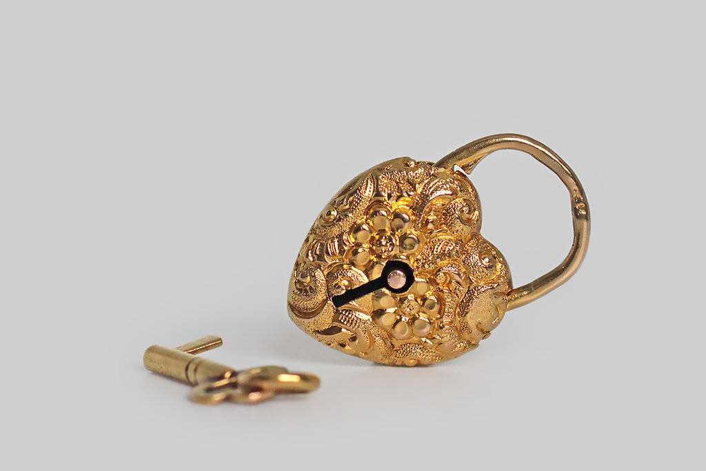 A lovely, miniature, Victorian-era, repoussé heart padlock, modeled in 14k yellow gold, and still partnered with its original 14k gold key. This functioning lock is in wonderful condition. It is highly-textural, decorated all-over with flowers and fine, swirling, baroque forms— a pair of blooms rest at the center of the pattern. This padlock would have originally been used as the focal piece for a bracelet or necklace