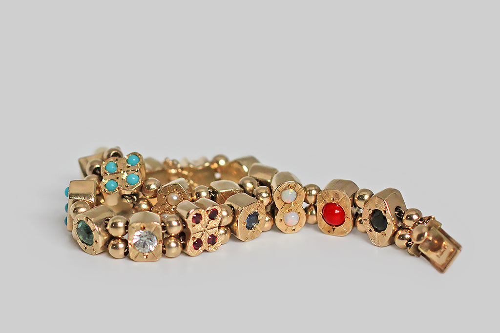 A charming 1940s slide charm bracelet, whose closely proportioned, gem-set charms are each a different geometric shape, and whose gemstones are placed like pips on card faces. These chunky little charms slide along the bracelet's two lengths of gold cable chain, separated by pairs of gold spacer beads— they feature a colorful variety of gemstones: sapphire, opal, turquoise glass, amethyst, blue and white zircon, coral, pearl, ruby and garnet. 