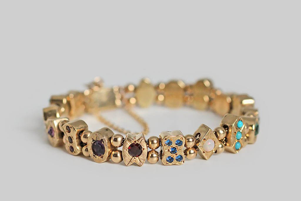 A charming 1940s slide charm bracelet, whose closely proportioned, gem-set charms are each a different geometric shape, and whose gemstones are placed like pips on card faces. These chunky little charms slide along the bracelet's two lengths of gold cable chain, separated by pairs of gold spacer beads— they feature a colorful variety of gemstones: sapphire, opal, turquoise glass, amethyst, blue and white zircon, coral, pearl, ruby and garnet. 