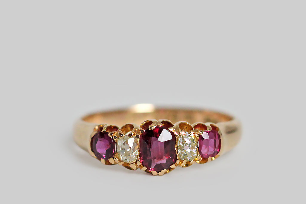 A darling, Victorian-era, five stone ring, modeled in 18k rosy yellow gold, with a tapering profile. This ring is set with three, vibrant, natural, rubies (.60 CTW) and two, cushion-shaped, old mine cut diamonds (.30 CTW)— these gemstones graduate in size, with the largest resting at the center. They are held in sculpted, split-prongs that are integral to the ring’s subtly-lobed, scalloped gallery. The ring's substaintial half-round shank tapers, somewhat, toward the base. 