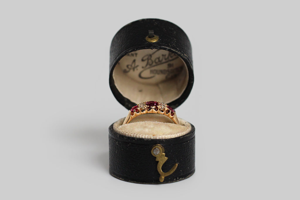 A darling, Victorian-era, five stone ring, modeled in 18k rosy yellow gold, with a tapering profile. This ring is set with three, vibrant, natural, rubies (.60 CTW) and two, cushion-shaped, old mine cut diamonds (.30 CTW)— these gemstones graduate in size, with the largest resting at the center. They are held in sculpted, split-prongs that are integral to the ring’s subtly-lobed, scalloped gallery. The ring's substaintial half-round shank tapers, somewhat, toward the base. 