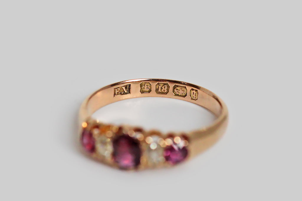 A darling, Victorian-era, five stone ring, modeled in 18k rosy yellow gold, with a tapering profile. This ring is set with three, vibrant, natural, rubies (.60 CTW) and two, cushion-shaped, old mine cut diamonds (.30 CTW)— these gemstones graduate in size, with the largest resting at the center. They are held in sculpted, split-prongs that are integral to the ring’s subtly-lobed, scalloped gallery. The ring's substaintial half-round shank tapers, somewhat, toward the base. 