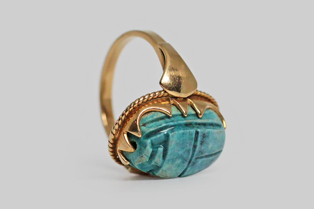 A magical, Art Deco era ring, modeled in 18k yellow gold and set with a brilliant blue, Egyptian, faience scarab bead. This talisman of renewal and rebirth is held in an ornately scalloped bezel. The foot of this festoon bezel is embellished with twisted gold wire, and the ring’s shoulders are decorated with a shapely, very minimal, lotus motif.
