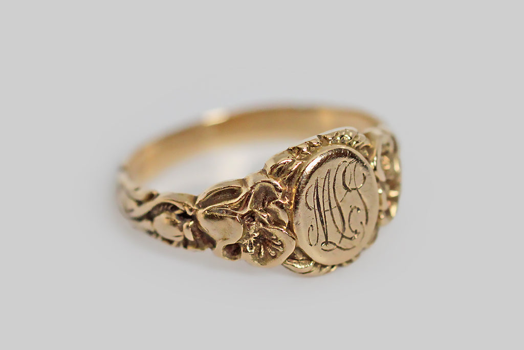 An antique, Art Nouveau era signet ring, made in 14k yellow gold, with ornate, carved details that are especially well-preserved. The ring’s shoulders are adorned with a pair of realistic, fleshy poppies, whose toothed leaves travel down the ring shank to the base, where their stems twine together. Its round face is engraved with the monogram MLJ, in curling period script. 